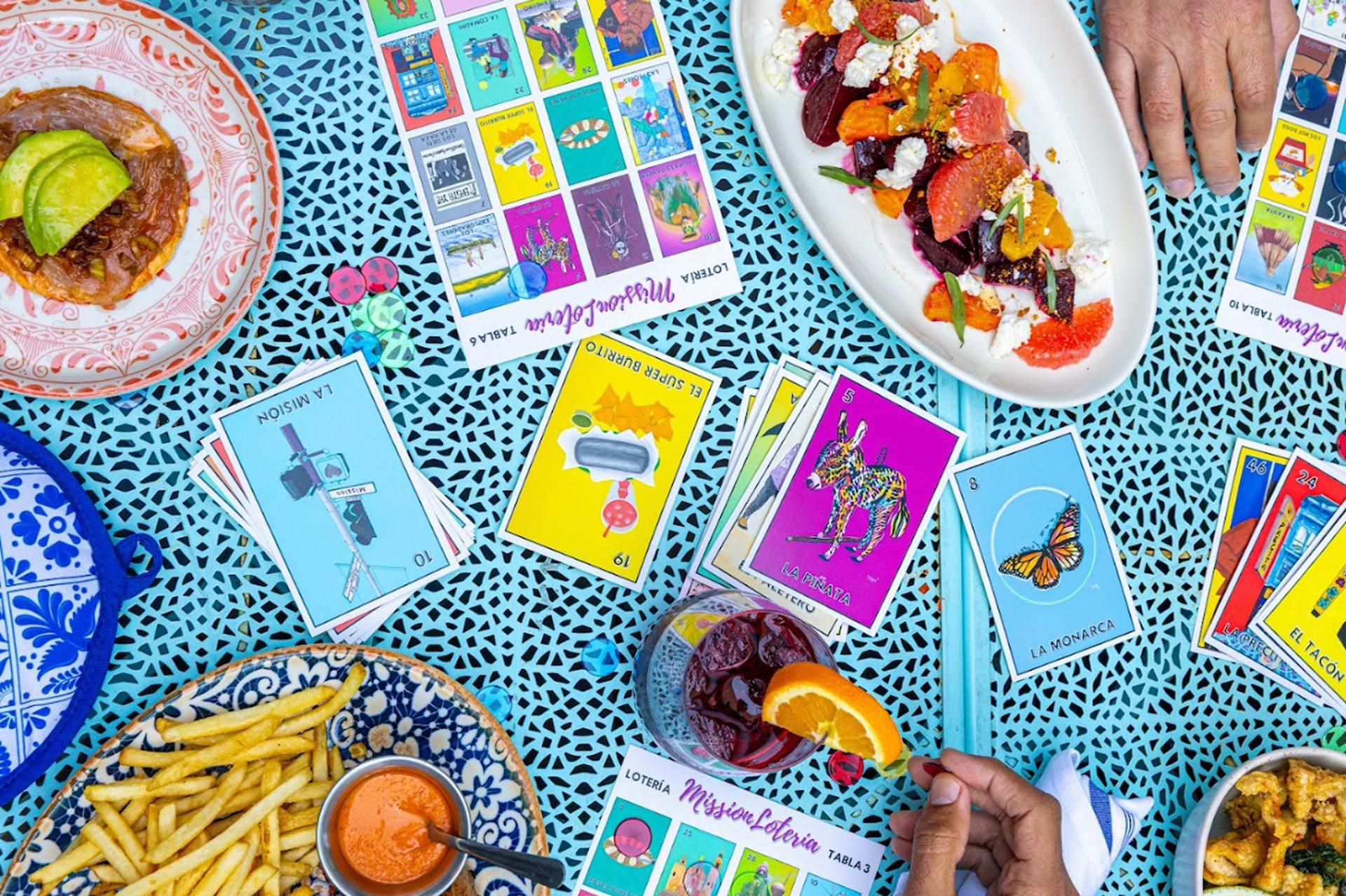 A Mission Lotería spread of food and lotería cards laid out on a table